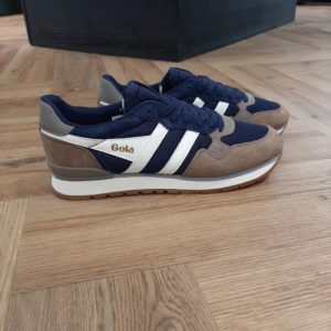 Gola Colorado Navy La sneaker Gola Colorado Navy est un classique réinventé issu des archives de la marque. Sa tige en maille polyester et daim est composée de bleu marine, de tabac et de blanc cassé. Cette chaussure est dotée d'une semelle intermédiaire en EVA et d'une semelle extérieure à crampons. C'est une basket incontournable pour hommes.  La marque Gola est une marque principalement connue pour ses chaussures de sport. Elle a été créé en 1905 à Leicester, au Royaume Uni. Pour l'anecdote, personne ne sait exactement d'où vient le nom de la marque. Pourtant, une légende urbaine raconte qu'elle doit son nom à l'anagramme du mot "Goal" (qui signifie "but" en anglais). La marque commence à être connue à la fin des années 1960. Notamment dans le sport notamment avec la Harrier qui reste aujourd'hui encore le modèle phare de la marque anglaise. En effet, elle a très vite été adoptée par le grand public. Fort de son histoire, la marque a su traverser les générations et est aujourd'hui une marque streetwear à part entière qui sait innover et suivre les tendances. On a d'ailleurs pu voir une collection 100 % vegan sortir des tuyaux en 2019, preuve de la capacité de Gola à s'adapter au marché actuel !