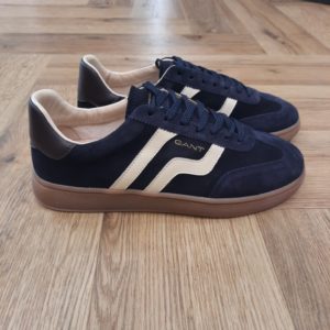 Gant Cuzmo Marine Les baskets Gant Cuzmo Marine sont fabriquées avec des matériaux de qualité supérieure qui reflètent l'essence du meilleur American Sportswear. Pour commencer, ce modèle se distingue par sa construction en daim doux, qui offre une finition élégante et durable. Puis, son design à silhouette basse, complété par une semelle en caoutchouc, garantit un ajustement confortable et sûr. Les semelles aux tons miel et les accents de couleurs contrastantes sur les côtés et à l'arrière ajoutent une touche moderne et distinctive. Elles sont idéales pour un look décontracté et urbain. En effet, ces baskets s'adaptent à différents styles, devenant une option polyvalente pour toute garde-robe. La marque Quand Bernard Gant, un immigrant ukrainien, arrive à New-York en 1914, il se plonge directement au cœur des métiers du prêt-à-porter. Il travaille dans un atelier de confection de chemises dans le centre de la ville. Ce n'est que quelques années plus tard, qu'il lancera l'entreprise familiale à l'aide de sa femme, également dans le métier. Rapidement, il fabrique des chemises pour les plus grandes marques du pays tel que Brooks Brothers. Si le style de Gant a beaucoup évolué au cours des années, il ne s'est jamais vraiment détaché de son image de marque Américaine traditionnelle. En effet, adepte du style Ivy avec comme produit phare la chemise à col boutonné. Après avoir été suédoise, la marque a été rachetée en 2008 par le groupe suisse Maus Frères, qui détient entre autres Lacoste et Aigle, mais elle garde encore ses valeurs originelles : celles de Bernard Gant.