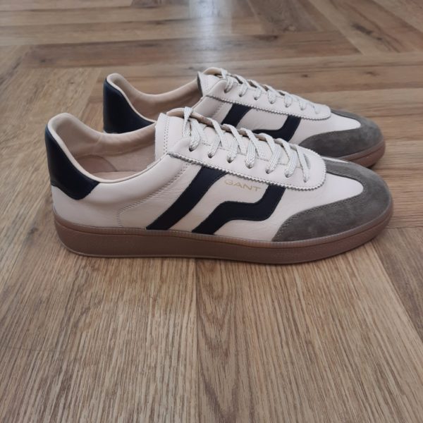 Gant Cuzmo Beige Les baskets Gant Cuzmo Beige sont fabriquées avec des matériaux de qualité supérieure qui reflètent l'essence du meilleur American Sportswear. Pour commencer, ce modèle se distingue par sa construction en daim doux, qui offre une finition élégante et durable. Puis, son design à silhouette basse, complété par une semelle en caoutchouc, garantit un ajustement confortable et sûr. Les semelles aux tons miel et les accents de couleurs contrastantes sur les côtés et à l'arrière ajoutent une touche moderne et distinctive. Elles sont idéales pour un look décontracté et urbain. En effet, ces baskets s'adaptent à différents styles, devenant une option polyvalente pour toute garde-robe. La marque Quand Bernard Gant, un immigrant ukrainien, arrive à New-York en 1914, il se plonge directement au cœur des métiers du prêt-à-porter. Il travaille dans un atelier de confection de chemises dans le centre de la ville. Ce n'est que quelques années plus tard, qu'il lancera l'entreprise familiale à l'aide de sa femme, également dans le métier. Rapidement, il fabrique des chemises pour les plus grandes marques du pays tel que Brooks Brothers. Si le style de Gant a beaucoup évolué au cours des années, il ne s'est jamais vraiment détaché de son image de marque Américaine traditionnelle. En effet, adepte du style Ivy avec comme produit phare la chemise à col boutonné. Après avoir été suédoise, la marque a été rachetée en 2008 par le groupe suisse Maus Frères, qui détient entre autres Lacoste et Aigle, mais elle garde encore ses valeurs originelles : celles de Bernard Gant.
