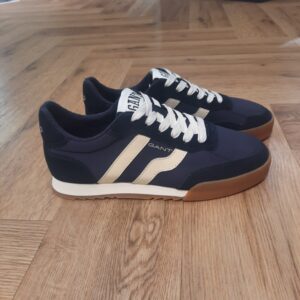 Gant Baylle Navy Les baskets Gant Baylle Navy sont la combinaison parfaite de style rétro et de confort. En effet, leur design intemporel et leurs matériaux haut de gamme en font un incontournable pour ceux qui cherchent à se démarquer avec un look élégant et sportif.  Les baskets Gant Baylle sont un modèle qui reflète l'essence du American Sportswear à travers leur construction en matériaux premium et un design intemporel. D'abord, leur tige texturée, élaborée en nubuck, est complétée par des détails en nylon et cuir. Celle-ci créée un équilibre parfait entre confort et style. Puis, les semelles en caoutchouc robustes et les découpes en EVA apportent une esthétique sportive inspirée du passé. Les chaussures idéales pour ceux qui souhaitent se démarquer avec un look vintage. Ces baskets ne sont pas seulement parfaites pour des activités décontractées. Mais, elles offrent également une excellente durabilité et un soutien au quotidien. Avec la basket Gant Baylle, chaque pas est léger et confortable, faisant de cette chaussure un choix essentiel pour toute garde-robe moderne. La marque Quand Bernard Gant, un immigrant ukrainien, arrive à New-York en 1914, il se plonge directement au cœur des métiers du prêt-à-porter. Il travaille dans un atelier de confection de chemises dans le centre de la ville. Ce n'est que quelques années plus tard, qu'il lancera l'entreprise familiale à l'aide de sa femme, également dans le métier. Rapidement, il fabrique des chemises pour les plus grandes marques du pays tel que Brooks Brothers. Si le style de Gant a beaucoup évolué au cours des années, il ne s'est jamais vraiment détaché de son image de marque Américaine traditionnelle. En effet, adepte du style Ivy avec comme produit phare la chemise à col boutonné. Après avoir été suédoise, la marque a été rachetée en 2008 par le groupe suisse Maus Frères, qui détient entre autres Lacoste et Aigle, mais elle garde encore ses valeurs originelles : celles de Bernard Gant