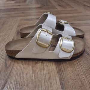 Arizona Big Buckle L’Arizona Big Buckle, classique BIRKENSTOCK au design intemporel, comble hommes et femmes depuis des décennies. Cette version s'enrichit de deux caractéristiques très appréciées : un revêtement discrètement satiné et une grande et élégante boucle ardillon. Sa tige a été confectionnée en Birko-Flor®, matériau synthétique doux et résistant.  Lit de pied anatomique en liège et latex  Tige : Birko-Flor®  Semelle de propreté : cuir velours  Semelle d’usure : EVA  Détails : deux brides réglables à grande boucle ardillon en métal  « Made in Germany »  N° d'art.1020021 L’histoire de Birkenstock Tout commence en 1774, lorsque Johann Adam Birkenstock, un cordonnier allemand passionné, fonde ce qui allait devenir l'une des marques de chaussures les plus emblématiques au monde. À l'époque, Johann menait une petite entreprise modeste qui lui permettait de vivre confortablement. Mais comment cette production familiale a-t-elle pu se transformer en un empire mondial de la chaussure ? Le tournant se produit en 1896 avec Konrad Birkenstock, un descendant de la famille. Konrad, visionnaire, décide de donner un coup de jeune à l'entreprise familiale. C'est lui qui a développé la première semelle intérieure orthopédique, une innovation qui a littéralement mis le sabot Birkenstock sur la carte mondiale. Cependant, il aura fallu plusieurs générations et une évolution constante pour que Birkenstock devienne la marque internationale que l'on connaît aujourd'hui. Le grand bond en avant s’est produit au XXe siècle, notamment aux États-Unis dans les années 1960, où les sandales Birkenstock sont devenues un symbole de la contre-culture et du confort. Mais ce n'est pas seulement l'histoire de l'entreprise qui fascine, c'est aussi sa philosophie. Birkenstock s'engage à produire des chaussures qui respectent la santé de vos pieds, en mettant l'accent sur la posture et le confort. La marque prône une fabrication durable et éthique, en utilisant des matériaux naturels et écologiques. Tout cela, combiné à un savoir-faire artisanal de qualité, souvent fabriqué à la main en Allemagne, fait de Birkenstock une marque unique en son genre.