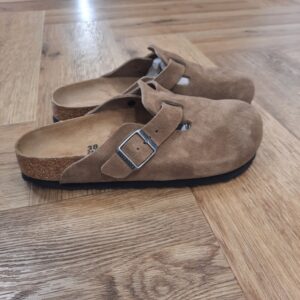 Boston Dark Tea Le sabot Boston Dark Tea, un classique parmi les modèles de BIRKENSTOCK, se présente ici dans un aspect moderne et la fois très naturel. C'est ce dont s'assure le dessus en cuir velours de grande qualité et particulièrement souple, au fini ciré tendance. L’histoire de Birkenstock Tout commence en 1774, lorsque Johann Adam Birkenstock, un cordonnier allemand passionné, fonde ce qui allait devenir l'une des marques de chaussures les plus emblématiques au monde. À l'époque, Johann menait une petite entreprise modeste qui lui permettait de vivre confortablement. Mais comment cette production familiale a-t-elle pu se transformer en un empire mondial de la chaussure ? Le tournant se produit en 1896 avec Konrad Birkenstock, un descendant de la famille. Konrad, visionnaire, décide de donner un coup de jeune à l'entreprise familiale. C'est lui qui a développé la première semelle intérieure orthopédique, une innovation qui a littéralement mis le sabot Birkenstock sur la carte mondiale. Cependant, il aura fallu plusieurs générations et une évolution constante pour que Birkenstock devienne la marque internationale que l'on connaît aujourd'hui. Le grand bond en avant s’est produit au XXe siècle, notamment aux États-Unis dans les années 1960, où les sandales Birkenstock sont devenues un symbole de la contre-culture et du confort.