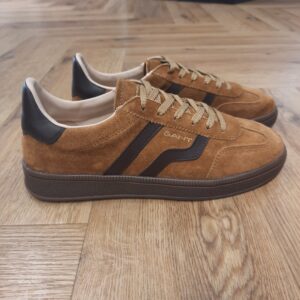 Cuzima Nut Brown Cuzima Nut Brown est un modèle d’inspiration rétro, qui se porte en toute saison. Tout d'abord, ces chaussures conçues avec une tige en daim lisse se distinguent par des finitions en cuir sur les côtés et au niveau du contrefort. Puis, la semelle extérieure en caoutchouc thermoplastique, associée à une doublure en tissu, confère un style vintage à l'ensemble. Les baskets Gant Cuzima pour femme sont la fusion parfaite entre style rétro et confort. Avec leur design intemporel et leurs matériaux de qualité supérieure, elles sont idéales pour toute occasion. Elles garantissent un look décontracté et moderne qui ne passera pas inaperçu. La marque Quand Bernard Gant, un immigrant ukrainien, arrive à New-York en 1914, il se plonge directement au cœur des métiers du prêt-à-porter. Il travaille dans un atelier de confection de chemises dans le centre de la ville. Ce n'est que quelques années plus tard, alors qu'il a déménagé à New Haven dans le Connecticut qu'il lancera l'entreprise familiale à l'aide de sa femme, également dans le métier. Rapidement, il fabrique des chemises pour les plus grandes marques du pays tel que Brooks Brothers. Si le style de Gant a beaucoup évolué au cours des années, il ne s'est jamais vraiment détaché de son image de marque Américaine traditionnelle. En effet, adepte du style Ivy avec comme produit phare la chemise à col boutonné. Après avoir été suédoise, la marque a été rachetée en 2008 par le groupe suisse Maus Frères, qui détient entre autres Lacoste et Aigle, mais elle garde encore ses valeurs originelles : celles de Bernard Gant.