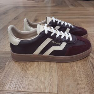 Cuzima Wine Cream Cuzima Wine Cream est un modèle d’inspiration rétro, qui se porte en toute saison. Tout d'abord, ces chaussures conçues avec une tige en cuir se distinguent par des finitions en daim lisse sur les côtés et au niveau du contrefort. Puis, la semelle extérieure en caoutchouc thermoplastique, associée à une doublure en tissu, confère un style vintage à l'ensemble. Les baskets Gant Cuzima pour femme sont la fusion parfaite entre style rétro et confort. Avec leur design intemporel et leurs matériaux de qualité supérieure, elles sont idéales pour toute occasion. Elles garantissent un look décontracté et moderne qui ne passera pas inaperçu. La marque Quand Bernard Gant, un immigrant ukrainien, arrive à New-York en 1914, il se plonge directement au cœur des métiers du prêt-à-porter. Il travaille dans un atelier de confection de chemises dans le centre de la ville. Ce n'est que quelques années plus tard, alors qu'il a déménagé à New Haven dans le Connecticut qu'il lancera l'entreprise familiale à l'aide de sa femme, également dans le métier. Rapidement, il fabrique des chemises pour les plus grandes marques du pays tel que Brooks Brothers. Si le style de Gant a beaucoup évolué au cours des années, il ne s'est jamais vraiment détaché de son image de marque Américaine traditionnelle. En effet, adepte du style Ivy avec comme produit phare la chemise à col boutonné. Après avoir été suédoise, la marque a été rachetée en 2008 par le groupe suisse Maus Frères, qui détient entre autres Lacoste et Aigle, mais elle garde encore ses valeurs originelles : celles de Bernard Gant.