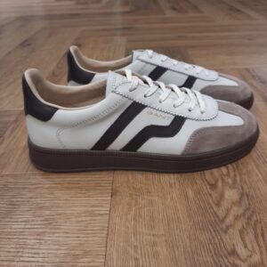 Cuzima Off White Brown Cuzima Off White Brown est un modèle d’inspiration rétro, qui se porte en toute saison. Tout d'abord, ces chaussures conçues avec une tige en cuir se distinguent par des finitions en daim lisse sur les côtés et au niveau du contrefort. Puis, la semelle extérieure en caoutchouc thermoplastique, associée à une doublure en tissu, confère un style vintage à l'ensemble. Les baskets Gant Cuzima pour femme sont la fusion parfaite entre style rétro et confort. Avec leur design intemporel et leurs matériaux de qualité supérieure, elles sont idéales pour toute occasion. Elles garantissent un look décontracté et moderne qui ne passera pas inaperçu. La marque Quand Bernard Gant, un immigrant ukrainien, arrive à New-York en 1914, il se plonge directement au cœur des métiers du prêt-à-porter. Il travaille dans un atelier de confection de chemises dans le centre de la ville. Ce n'est que quelques années plus tard, alors qu'il a déménagé à New Haven dans le Connecticut qu'il lancera l'entreprise familiale à l'aide de sa femme, également dans le métier. Rapidement, il fabrique des chemises pour les plus grandes marques du pays tel que Brooks Brothers. Si le style de Gant a beaucoup évolué au cours des années, il ne s'est jamais vraiment détaché de son image de marque Américaine traditionnelle. En effet, adepte du style Ivy avec comme produit phare la chemise à col boutonné. Après avoir été suédoise, la marque a été rachetée en 2008 par le groupe suisse Maus Frères, qui détient entre autres Lacoste et Aigle, mais elle garde encore ses valeurs originelles : celles de Bernard Gant.
